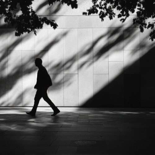 Silhouette of a person walking along a paved sidewalk beside a building wall, with tree shadows and sunlight creating patterns on the surface.