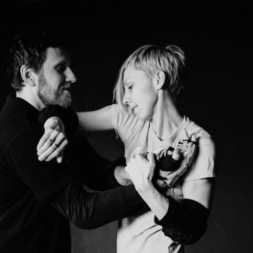 Two people practicing martial arts in close contact, one using an arm lock technique on the other in a controlled manner, set against a dark background.