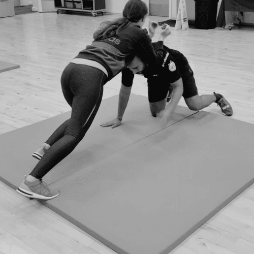 Two people engage in a grappling exercise on a mat in a gym setting.
