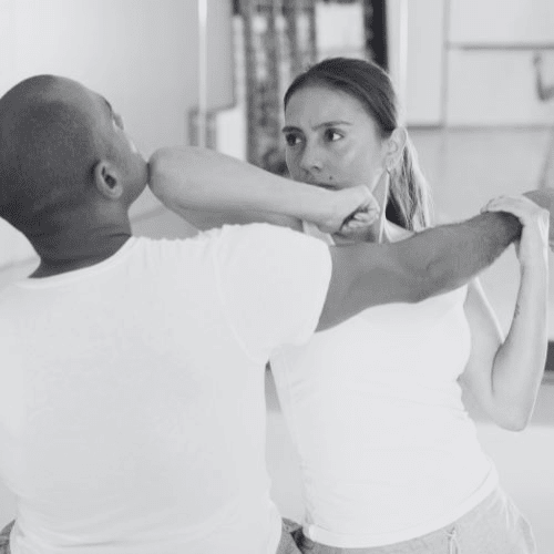 Two people, one woman, practicing martial arts in a room, one blocking a punch with an elbow. Both wearing white tops.
